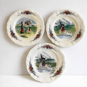 Peut inclure: Trois assiettes décoratives vintage avec des bords festonnés. Chaque assiette présente une scène de paysage centrale avec des personnages et une bordure florale rouge, bleue et jaune. Les assiettes sont blanc cassé.