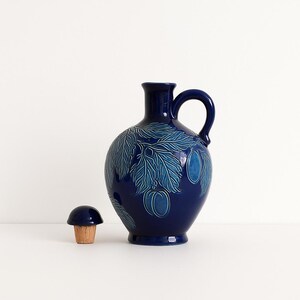 Peut inclure: Une cruche en céramique bleu foncé avec une anse et un bouchon assorti. La cruche est ornée d'un motif de feuilles et de fruits dans des tons de bleu. Le bouchon est en forme de champignon.