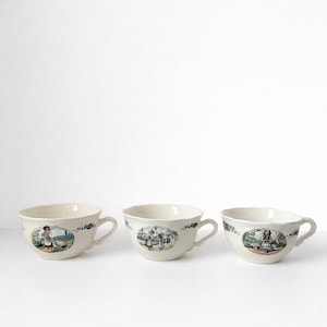 Peut inclure: Trois tasses à thé en céramique blanc cassé avec des anses. Chaque tasse présente une scène pastorale différente dans un cadre circulaire. Les scènes incluent des personnages en costume d'époque. Des motifs floraux ornent les bords des tasses.