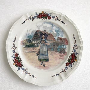 Puede incluir: Un plato decorativo blanco con borde festoneado. El plato presenta una escena pintada de una mujer con vestimenta tradicional frente a una casa. Diseños florales y de vid en rojo y azul adornan el borde.