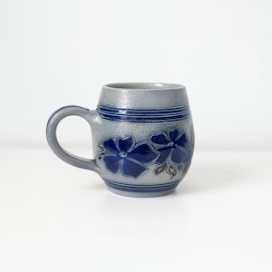Peut inclure: Mug en céramique grise avec une anse, orné de motifs floraux bleus. La tasse présente des bandes bleues en haut et en bas. Une pièce classique et décorative pour les boissons.