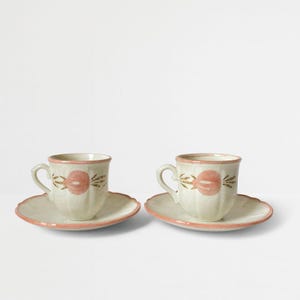 Könnte beinhalten: Zwei Keramik-Teetassen und Untertassen. Die Tassen und Untertassen sind cremefarben mit einem rosa Blumendesign. Die Ränder der Untertassen sind ebenfalls rosa. Die Tassen haben einen Henkel und stehen auf einer weißen Oberfläche.