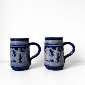 Peut inclure: Deux chopes à bière en céramique bleue avec des sculptures en relief grises de personnes dansant. Chaque chope a une anse bleu foncé et un bord gris. Les chopes sont sur une surface blanche.
