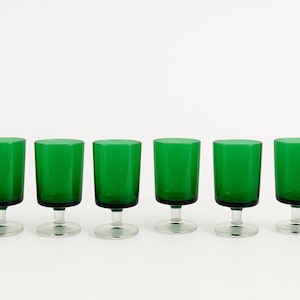 Peut inclure: Six gobelets en verre teinté vert, avec un bol court et cylindrique et une tige et une base transparentes. Les gobelets sont disposés en rangée sur fond blanc. La verrerie est probablement destinée à servir des boissons.