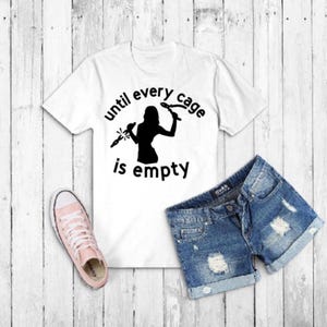 May include: White t-shirt with the text "until every cage is empty" and a black silhouette graphic. The image also includes denim shorts and a pair of pink sneakers.