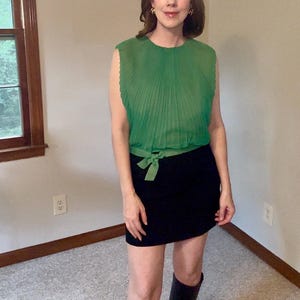 May include: A sleeveless green blouse featuring a pleated texture and a matching fabric tie at the waist, paired with a solid black skirt. The blouse has a round neckline and a semi-sheer appearance, while the skirt is a simple, straight-cut design.