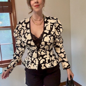 May include: A long-sleeved blazer featuring a bold black and white abstract pattern, a deep V-neckline, and a peplum-style hem. The garment is worn over a black lace-trimmed camisole and paired with black shorts, creating a structured silhouette.