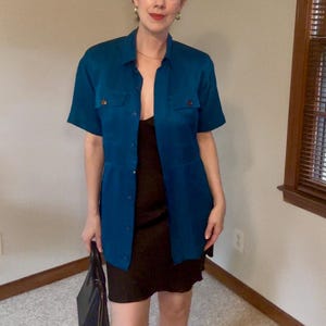 May include: A short-sleeved teal button-down shirt with two chest pockets worn over a black slip dress. The shirt features a collar and a button-front closure. A black handbag is held at the side. The ensemble is layered for a coordinated look.