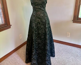 May include: A strapless, floor-length formal dress in dark green with a floral pattern. The dress features a fitted bodice and a full skirt. The top of the bodice is embellished with a subtle, sparkling trim. The dress is displayed on a mannequin.