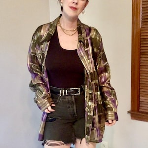 May include: A long-sleeve button-down shirt featuring a metallic gold, purple, and dark green abstract block pattern. The shirt is worn over a solid black scoop-neck tank top, paired with black denim shorts that have a distressed, frayed hemline.