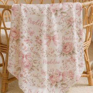 May include: A baby blanket with a floral and bow pattern in shades of pink and green, with the name "Madeline" printed across the blanket. The blanket is draped over a wicker bassinet.