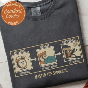 May include: Dark gray t-shirt with a humorous graphic illustrating the sequence of events: alarm rings, snooze button, and being late for work. The text "MASTER THE SEQUENCE" is below the graphic. A "Comfort Colors" brand logo is in the upper left corner.