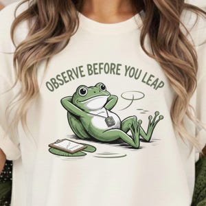 May include: Cream-colored t-shirt featuring a relaxed green frog illustration with the text "OBSERVE BEFORE YOU LEAP". The frog is reclining with a clipboard and a thought bubble. The design is printed on a soft, casual tee.