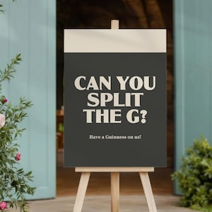 以下が含まれることがあります： 木製のイーゼルが、白い文字で「CAN YOU SPLIT THE G？」と書かれた看板を支えています。背景は濃いグレーです。その下には「Have a Guinness on us！」と書かれています。看板は、ドアと緑の背景の前に置かれています。