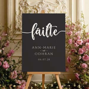 Può includere: Un cartello nero su un cavalletto di legno con testo bianco che recita "fáilte", "ANN-MARIE e ODHRAN" e "06.07.28". Il cartello è circondato da fiori rosa e verde.