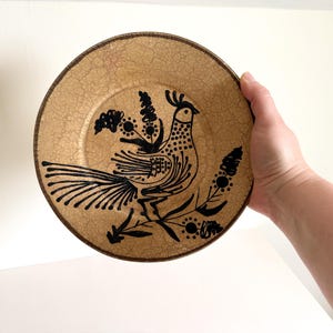 May include: A decorative plate with a tan background and a black bird design. The bird is surrounded by floral elements. The plate has a textured surface and a dark brown rim. The plate is round.