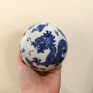 Puede incluir: Una esfera de cerámica blanca con un diseño de dragón azul. El dragón está intrincadamente detallado, con escamas y una melena fluida. La esfera también presenta motivos de nubes azules.