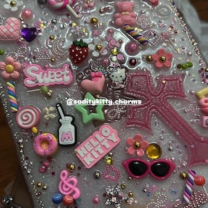 Könnte beinhalten: Eine transparente Handyhülle, verziert mit verschiedenen rosa und weißen Charms, darunter Hello Kitty, Lollis, Herzen und das Wort "Sweet". Die Hülle ist mit Glitzer und kleinen Perlen verziert und schafft eine verspielte und mädchenhafte Ästhetik.