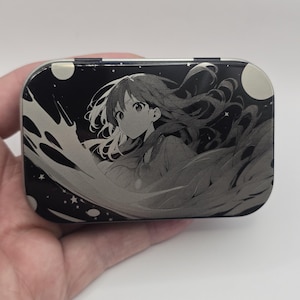 Op de afbeelding: Een rechthoekige, zwart-witte blikken doos met een anime-achtige illustratie van een meisje met lang haar. Het ontwerp bevat sterren en een maan, wat een hemels thema creëert. De doos wordt in een hand gehouden.