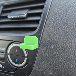 Könnte beinhalten: Ein hellgrüner, 3D-gedruckter Handyhalter für die Lüftung im Auto. Der Halter ist rechteckig mit einer gebogenen Oberseite und an einem schwarzen Armaturenbrett befestigt. Das Armaturenbrett hat Lüftungsschlitze, Knöpfe und eine strukturierte Oberfläche.