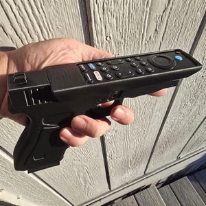 May include: A black television remote control integrated into a handgun-shaped housing. The remote features a circular navigation pad, volume and channel controls, and dedicated buttons for Netflix, Peacock, and Prime Video, with a blue power button.