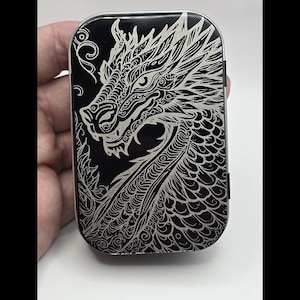 Puede incluir: Un estuche rectangular de metal negro con un diseño detallado de dragón plateado. El dragón está intrincadamente grabado, mostrando escamas, una expresión feroz y patrones arremolinados. El estuche tiene bordes redondeados y un acabado brillante.