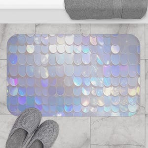 May include: A rectangular bath mat with an iridescent, scale-like design in shades of white, blue, and purple. The mat is placed on a tiled surface, with a pair of gray knit slippers at the bottom. The mat's design resembles fish scales.