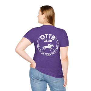 May include: Purple t-shirt with a white circular graphic on the back. The graphic features the text "OTT Club" above a horse and rider, with "Race + Retire + Restart" around the circle. The shirt is worn by a person in blue jeans.