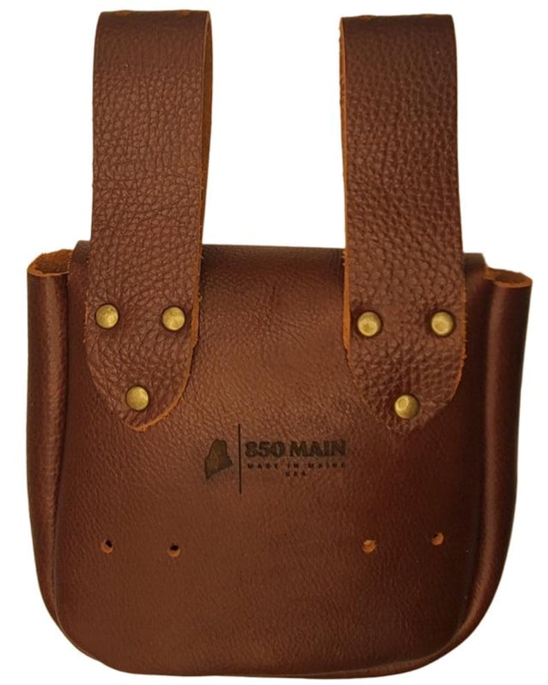 Op de afbeelding: Een bruin leren tasje met twee verticale bandjes en messing klinknagels. Het tasje heeft een gebogen bodem en de tekst "850 MAIN MADE IN MAINE USA" is zichtbaar op de voorkant.