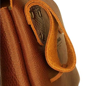 Op de afbeelding: Close-up van een bruine leren tas met een lusvormige handgreep. De tas heeft een getextureerd oppervlak en is afgewerkt met lichtere bruine leren bekleding en metalen klinknagels. De handgreep is omgevouwen en vastgezet met een leren riem.
