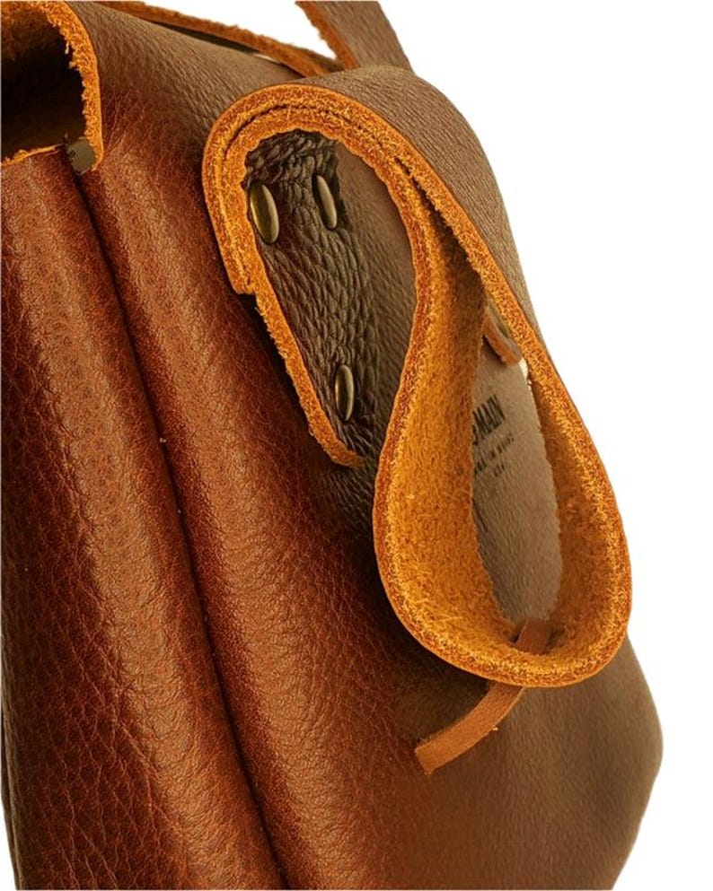 Op de afbeelding: Close-up van een bruine leren tas met een lusvormige handgreep. De tas heeft een getextureerd oppervlak en is afgewerkt met lichtere bruine leren bekleding en metalen klinknagels. De handgreep is omgevouwen en vastgezet met een leren riem.
