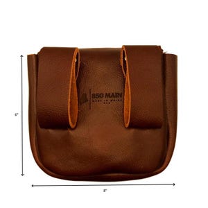 Op de afbeelding: Een bruine leren tas, ongeveer 20 cm breed en 15 cm hoog, met een gebogen bodem en twee lussen aan de achterkant. De tekst "850 MAIN MADE IN MAINE USA" is op de voorkant gedrukt.