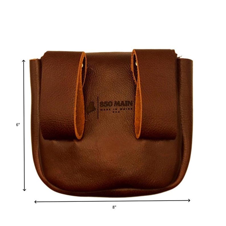 Op de afbeelding: Een bruine leren tas, ongeveer 20 cm breed en 15 cm hoog, met een gebogen bodem en twee lussen aan de achterkant. De tekst "850 MAIN MADE IN MAINE USA" is op de voorkant gedrukt.