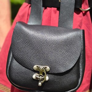 Op de afbeelding: Een zwarte leren tas met een klepsluiting en een zilverkleurige sluiting. De tas is bevestigd aan een zwarte riem met twee riemen. De achtergrond is een rood kledingstuk.