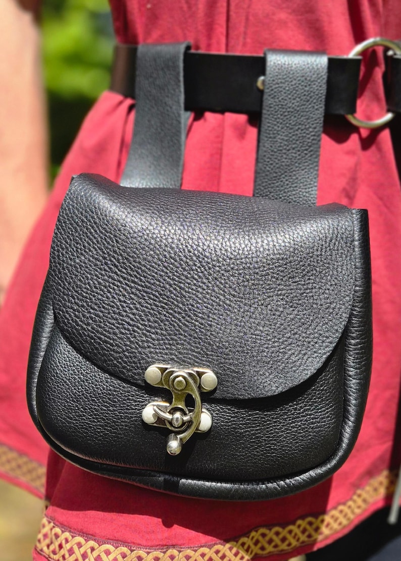 Op de afbeelding: Een zwarte leren tas met een klepsluiting en een zilverkleurige sluiting. De tas is bevestigd aan een zwarte riem met twee riemen. De achtergrond is een rood kledingstuk.