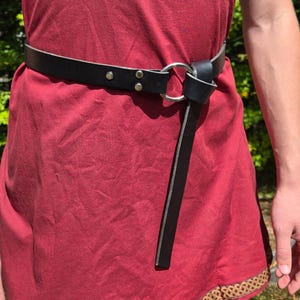 Può includere: Cintura in pelle nera con chiusura ad anello in metallo argentato. La cintura è indossata sopra un abito rosso. La cintura ha due rivetti argentati vicino alla fibbia. La cintura è un design semplice.