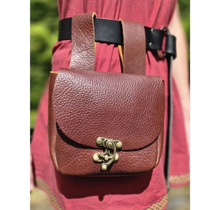Op de afbeelding: Een bruine leren tas met een getextureerd oppervlak, bevestigd aan een zwarte riem. De tas heeft een gebogen klep en een decoratieve metalen sluiting. De riem wordt om de taille gedragen.