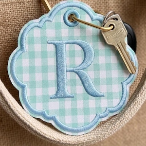 May include: A scalloped, light blue and white gingham fabric keyring featuring a large, embroidered blue letter R in the centre. The keyring is attached to a gold-toned metal ring that holds a silver-coloured metal key with the word Master.