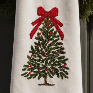 May include: A white kitchen towel featuring an embroidered green Christmas tree with a textured brown trunk. The tree is decorated with small red embroidered dots and topped with a large, detailed red bow stitched in a cross-hatch pattern.