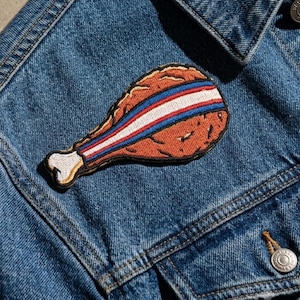 Könnte beinhalten: Ein gestickter Aufnäher in Form einer frittierten Hähnchenkeule mit goldbrauner, strukturierter Oberfläche, einem weißen Knochenende und einem dekorativen diagonalen Streifen in Rot, Weiß und Blau, befestigt auf blauem Jeansstoff.