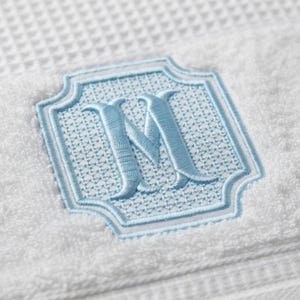 Puede incluir: Una toalla blanca de tejido de gofre con un monograma bordado en azul claro de la letra M. La letra está centrada dentro de un marco de cuatrifolio cosido en azul claro, relleno con un patrón de trama cruzada de hilo azul claro.