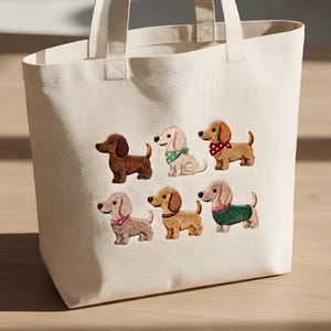 May include: A cream-colored canvas tote bag featuring an embroidered design of six dachshund dogs. The dogs are depicted in various shades of brown and cream, wearing accessories including a green polka-dot bandana, a red polka-dot bandana, a pink bow, and a green sweater.