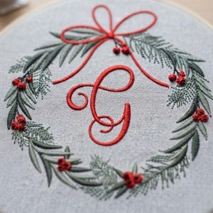 以下が含まれることがあります： 薄いグレーの布地を張った円形の刺繍枠。手縫いのリース模様が施されており、緑の松葉と赤い実が描かれています。上部には赤いリボンの結び目があり、中央には大きな赤い筆記体の「G」の文字が刺繍されています。
