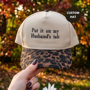 May include: A beige baseball cap with a leopard print brim. The front of the cap features black embroidered text that reads, "Put it on my Husband's tab." The cap has a structured crown and a button on top, with a dark circular sticker.