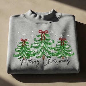 May include: A light gray mock neck sweatshirt featuring an embroidered design of three green pine trees with brown trunks. Each tree is topped with a red bow. White embroidered snowflakes surround the trees, and the text Merry Christmas is stitched in black cursive script below.