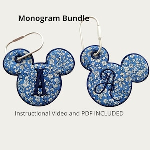 May include: Two mouse-shaped fabric keychains featuring a blue floral pattern with dark blue embroidered monograms of the letter A in different fonts. Each keychain includes a silver metal ring and the text Monogram Bundle and Instructional Video and PDF INCLUDED.