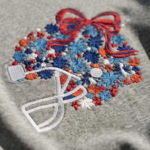 Puede incluir: Una sudadera gris jaspeado con un diseño bordado de una regadera blanca rebosante de un denso grupo de pequeñas flores azules, naranjas y blancas, rematado con un gran lazo rojo con contornos cosidos en azul oscuro.
