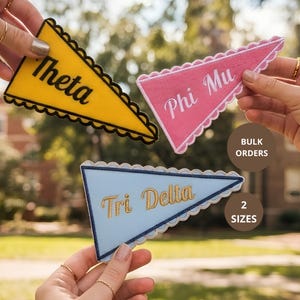 May include: Three triangular pennant patches with scalloped edges. One is yellow with black text reading Theta, one is pink with white text reading Phi Mu, and one is light blue with gold text reading Tri Delta. Text on image reads BULK ORDERS 2 SIZES.