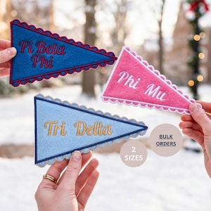 May include: Three triangular felt pennants with scalloped edges. The blue pennant features red embroidered text reading Pi Beta Phi. The pink pennant has white embroidered text reading Phi Mu. The light blue pennant has gold embroidered text reading Tri Delta.