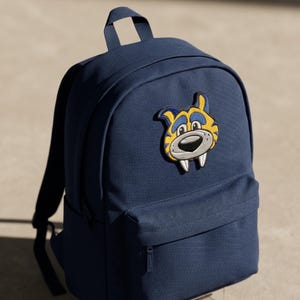 May include: A navy blue backpack featuring a large embroidered patch of a cartoon tiger head with yellow and blue fur, a grey snout, and two prominent white fangs. The backpack has a main zippered compartment, a front pocket, and a top carry handle.